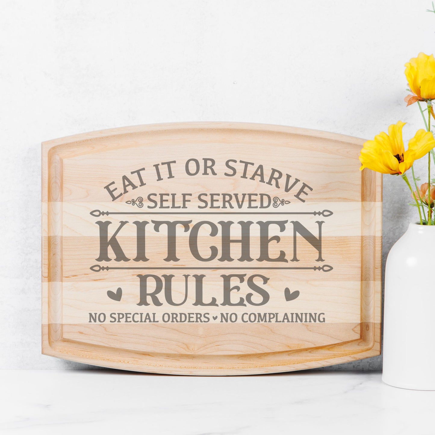 Arched maple cutting board against a white background with yellow flowers in a vase to the right of the cutting board