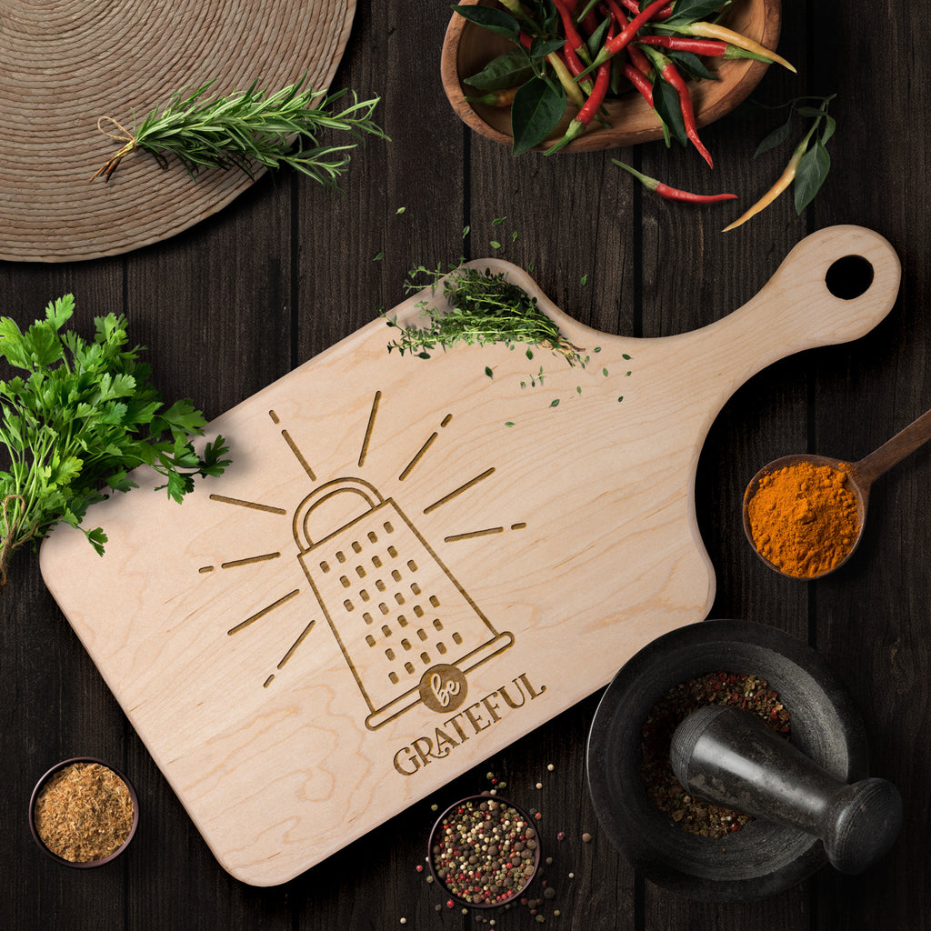 a maple cutting board with a cheese grater with the words "be grateful" engraved on the cutting board on a brown table with herb and spices as decoration.