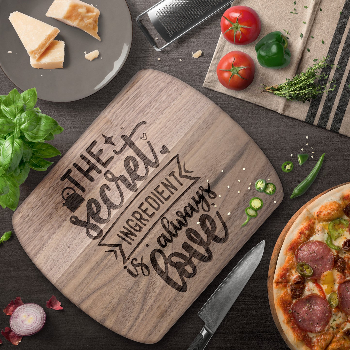 view of walnut cutting board with the words "the secret ingredient is always love" on a brown table with pizza, knife, tomatoes, and cheese as decoration