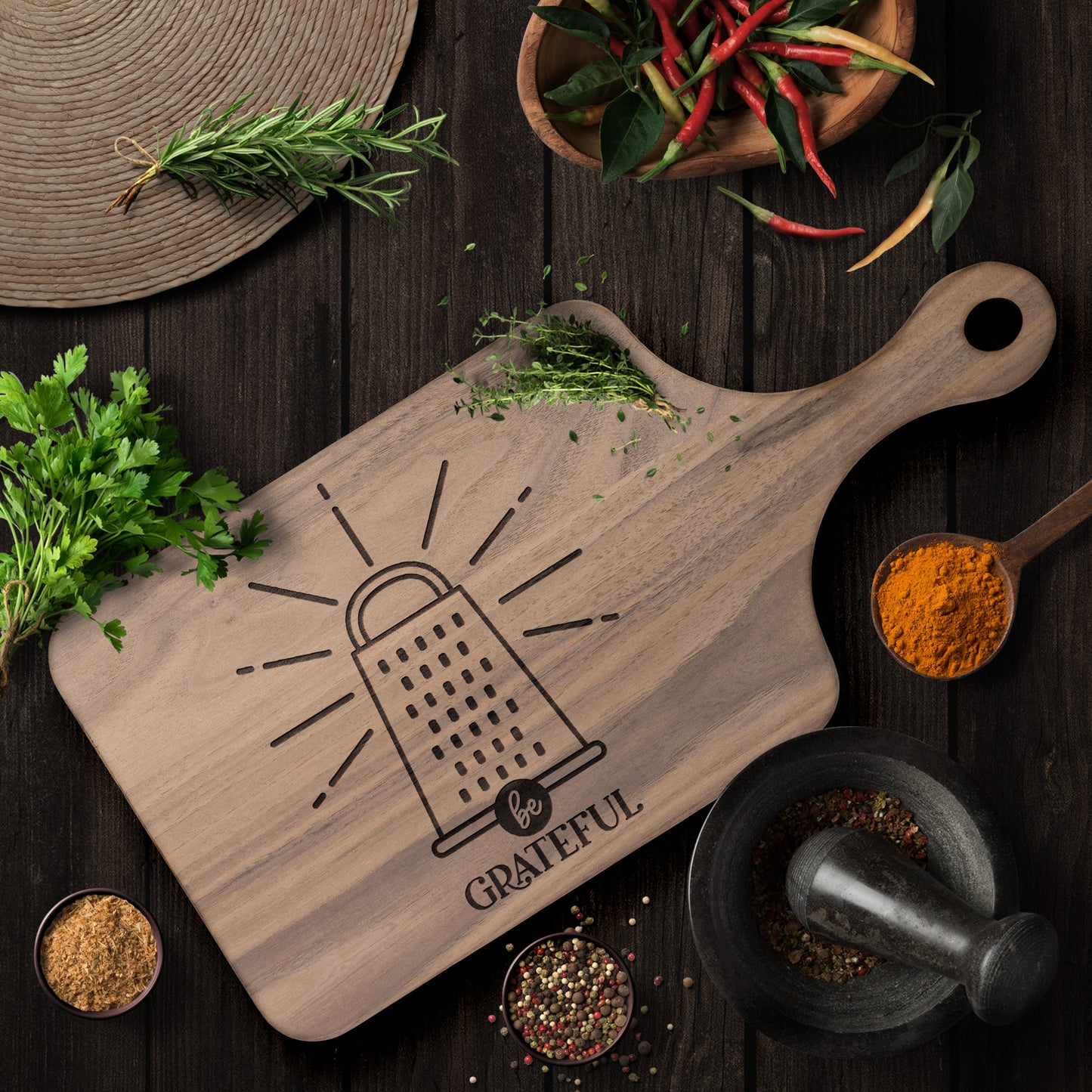 a walnut cutting board with a cheese grater with the words "be grateful" engraved on the cutting board on a brown table with herbs and spices as decoration