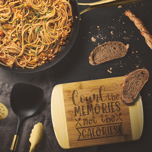 bamboo cutting board with the words "Count the Memories not the calories" on  a black table with paste and bread as decor