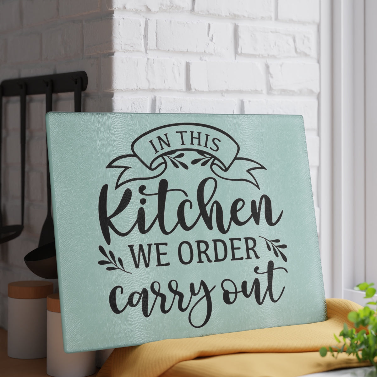 image of a glass cutting board with the words "in this kitchen we order carry out" leaning against a white brick wall