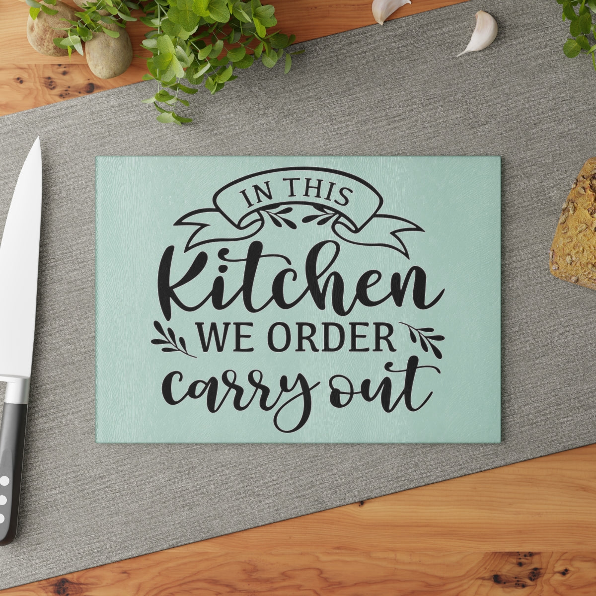 image of a glass cutting board with the words "in this kitchen we order carry out" on a brown table with a gray runner and knife as decoration